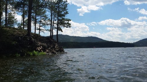 A serene lake scene with trees on the shore, mountains in the background, and a partly cloudy sky.