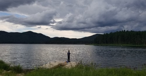 A person stands on a dock by a lake, gazing at the water under a cloudy sky with mountains in the background.