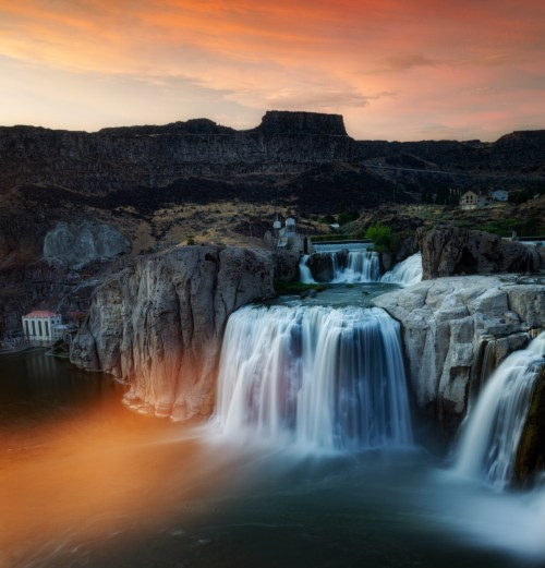 A stunning waterfall cascades over rocky cliffs at sunset, surrounded by rugged terrain and a serene river.