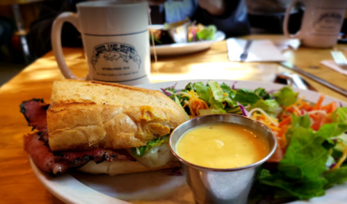 A sandwich with meat and lettuce, served with a side salad and a small cup of sauce, on a wooden table.