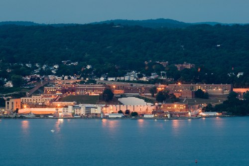 A nighttime view of a waterfront area with buildings and greenery in the background, reflecting lights on the water.