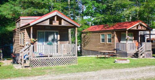 Two wooden cabins with red roofs in a wooded area, featuring porches and a picnic table in front.