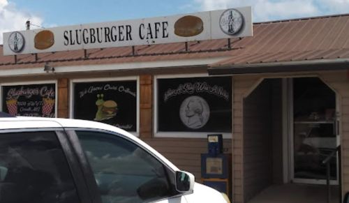 Exterior of Slugburger Café with a sign and windows displaying menu items, parked white vehicle in foreground.