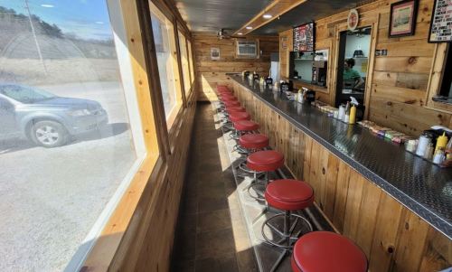 A rustic diner interior with red stools at a long counter, large windows, and a view of a car outside.