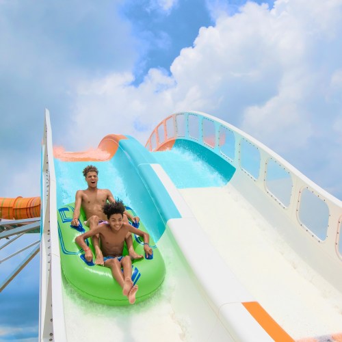 Two boys slide down a colorful water slide, splashing into a pool under a bright blue sky.