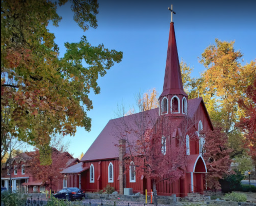 A red church with a tall steeple surrounded by colorful autumn trees and a parked car in the foreground.