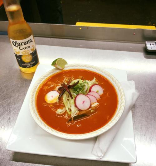 A bowl of red soup garnished with radishes and lettuce, alongside a bottle of Corona beer and a lime wedge.