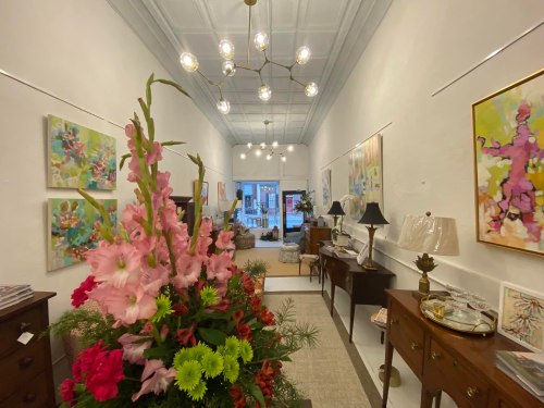 A bright gallery interior featuring colorful artwork, floral arrangements, and vintage furniture.