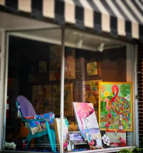 A colorful art studio window display featuring paintings, a chair, and decorative flamingo items.