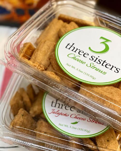 Two clear containers of cheese straws labeled "Three Sisters" and "Jalapeño Cheese Straws," showcasing golden-brown snacks.