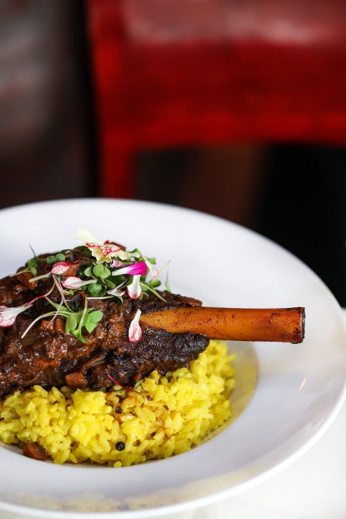 A plate of tender lamb shank on a bed of yellow rice, garnished with microgreens and edible flowers.