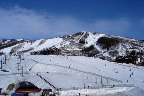 Ski resort scene with slopes, skiers, and a chairlift against a backdrop of snow-covered mountains and blue sky.