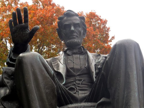 Statue of Abraham Lincoln seated, raising his right hand, surrounded by autumn foliage.