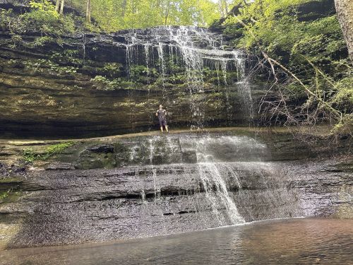 A person stands near a waterfall cascading over rocky cliffs, surrounded by lush greenery and a calm pool below.