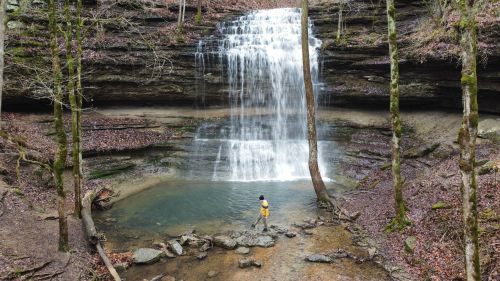 A person stands in shallow water near a waterfall surrounded by rocky terrain and trees in a serene forest setting.
