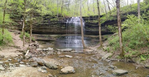 A serene waterfall cascades over a rocky ledge, surrounded by lush green trees and a calm stream below.