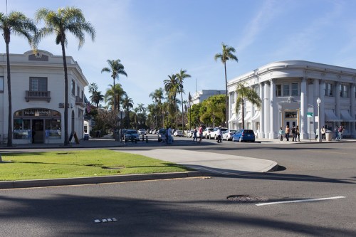 A sunny street scene with palm trees, featuring buildings and people walking near a crosswalk.