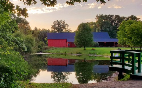 A serene landscape featuring a red barn reflected in a calm pond, surrounded by lush greenery and a wooden bridge.