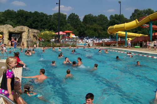 A busy outdoor pool with children and adults swimming, water slides, and colorful umbrellas in the background.
