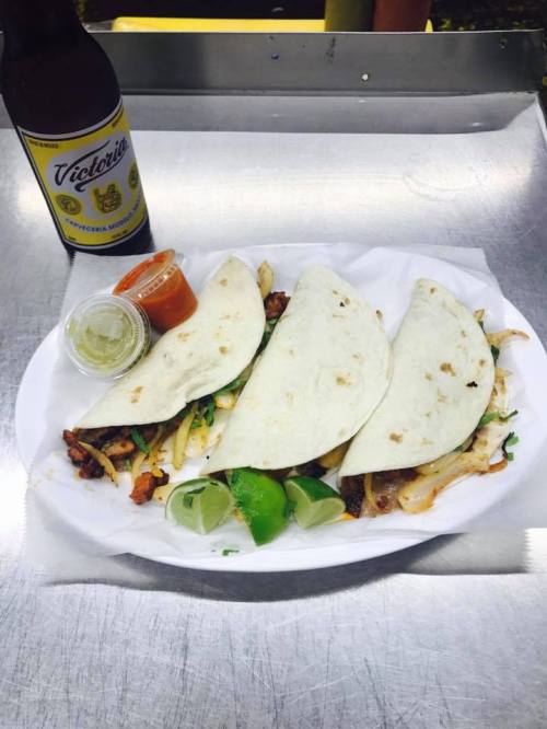 Three tacos on a plate with lime wedges, salsa, and a bottle of Victoria beer beside them.