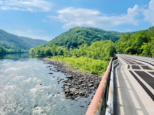 A scenic view of a river bordered by lush green hills, with a pathway running alongside the water under a clear blue sky.