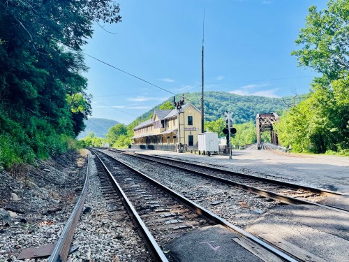 A scenic view of a railway track leading to a historic train station surrounded by lush greenery and mountains.