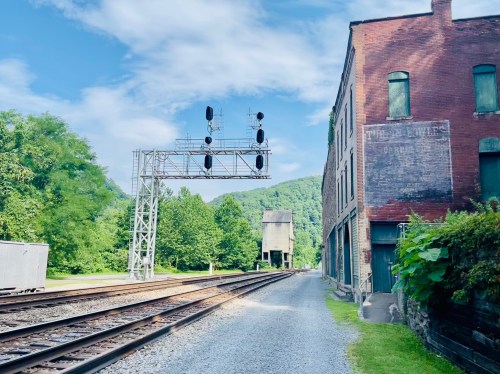 A railway track runs alongside a brick building, with a signal tower and green hills in the background under a blue sky.