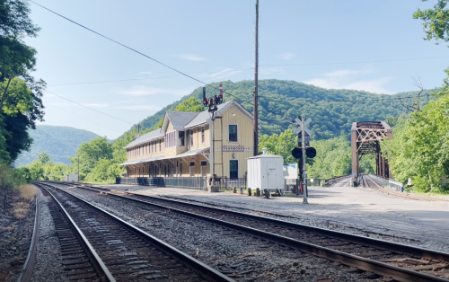 A vintage train station beside railway tracks, with a mountain backdrop and a signal post in the foreground.