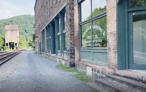 Abandoned building with large windows, gravel path, and railroad tracks beside a green hillside.