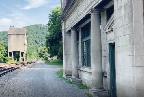 A historic building with columns beside a gravel path and railroad tracks, surrounded by greenery and hills.
