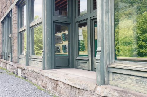 A stone building with large windows and a wooden entrance, surrounded by greenery and gravel.