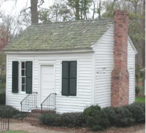 A small white house with a sloped roof, green shutters, and a brick chimney, surrounded by shrubs and trees.