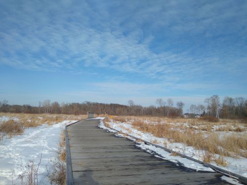 A wooden boardwalk leads through a snowy landscape under a blue sky with scattered clouds.
