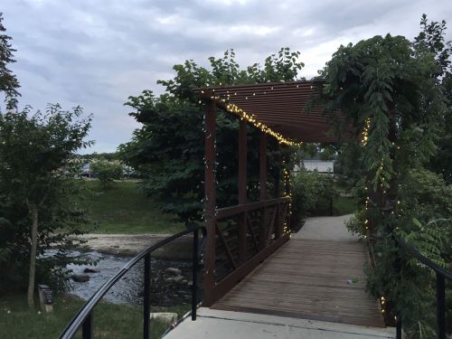 A wooden bridge adorned with string lights, surrounded by greenery and a stream under a cloudy sky.