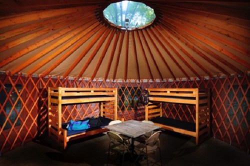 Interior of a yurt featuring wooden bunk beds, a table, and a skylight, surrounded by a lattice wall.