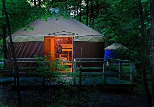 A cozy yurt in a forest, illuminated from within, with a wooden deck and surrounding greenery.