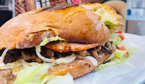 A close-up of a sandwich with grilled meat, lettuce, tomato, and a toasted bun, served on a white paper wrapper.