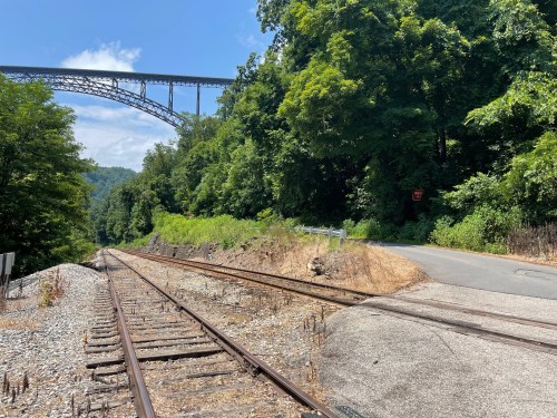 A view of a railway track beside a road, with a bridge and lush greenery in the background under a clear blue sky.