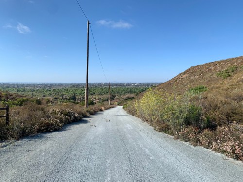 A gravel road winds through a hilly landscape, with power lines and a clear blue sky in the background.