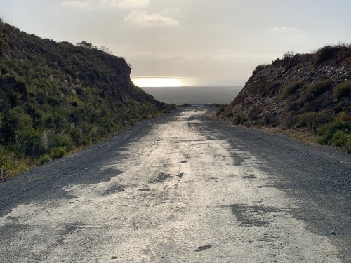 A winding, unpaved road between grassy hills, leading towards a distant view of the ocean under a cloudy sky.