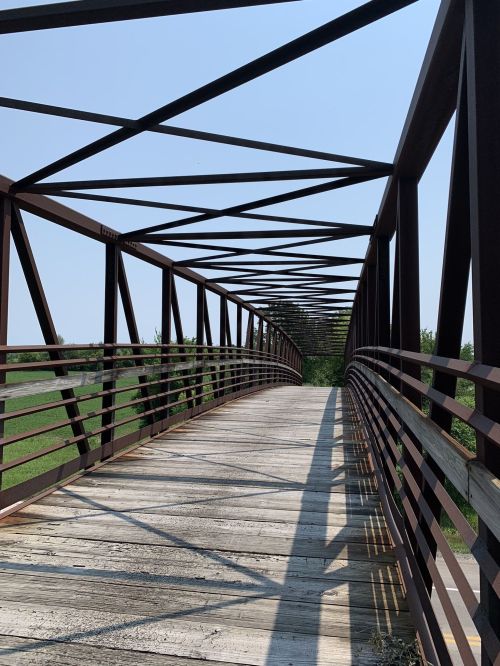 A metal bridge with a wooden walkway, surrounded by greenery under a clear blue sky. Shadows cast on the path.