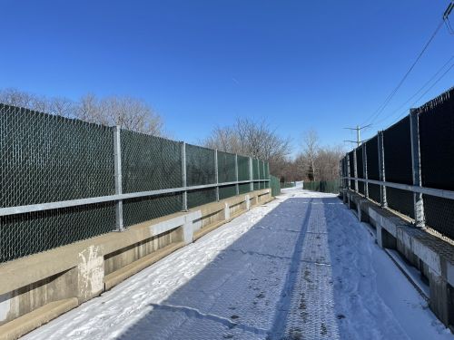 A snow-covered pathway flanked by green chain-link fences under a clear blue sky.