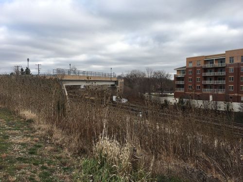 A view of a railway track beside a grassy area, with an overpass and an apartment building in the background under cloudy skies.