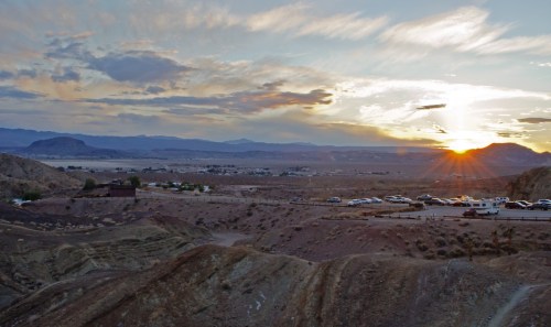 A scenic view of a desert landscape at sunset, with mountains in the background and parked vehicles in the foreground.