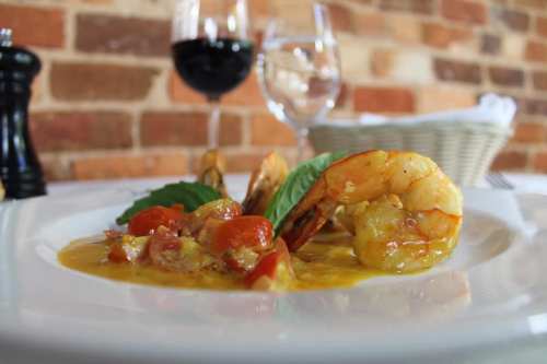 A plate of shrimp in a savory sauce, garnished with basil, with wine glasses and a basket in the background.