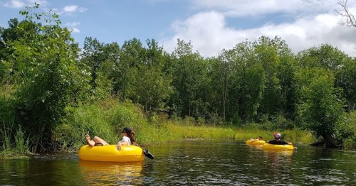Two people relax on yellow inner tubes in a calm river surrounded by lush greenery and trees under a blue sky.
