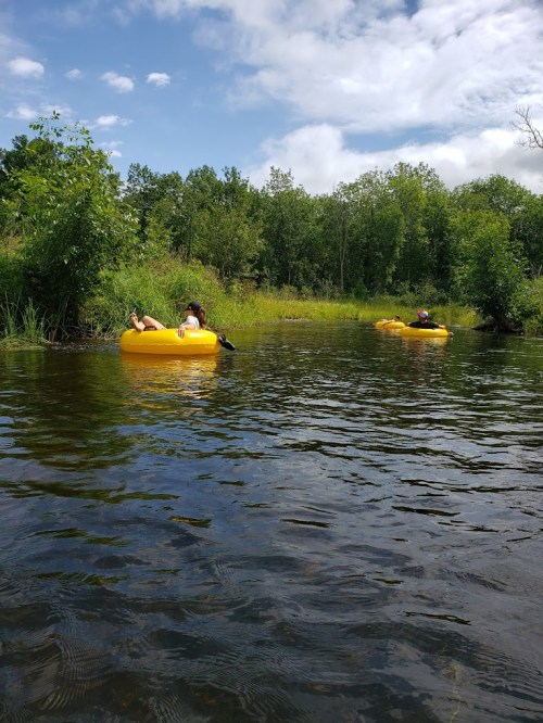 Two people relax on yellow inner tubes in a calm, green river surrounded by trees and a blue sky.