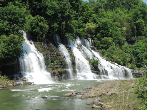 A scenic waterfall cascading over rocks, surrounded by lush green trees and a calm river below.