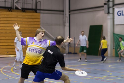 A player in a yellow and purple jersey gestures while another player prepares to throw a frisbee in a gymnasium.