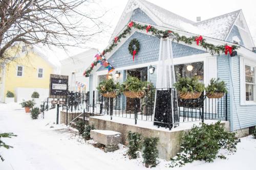 A cozy blue house decorated for the holidays, surrounded by snow and festive greenery, with a welcoming porch.
